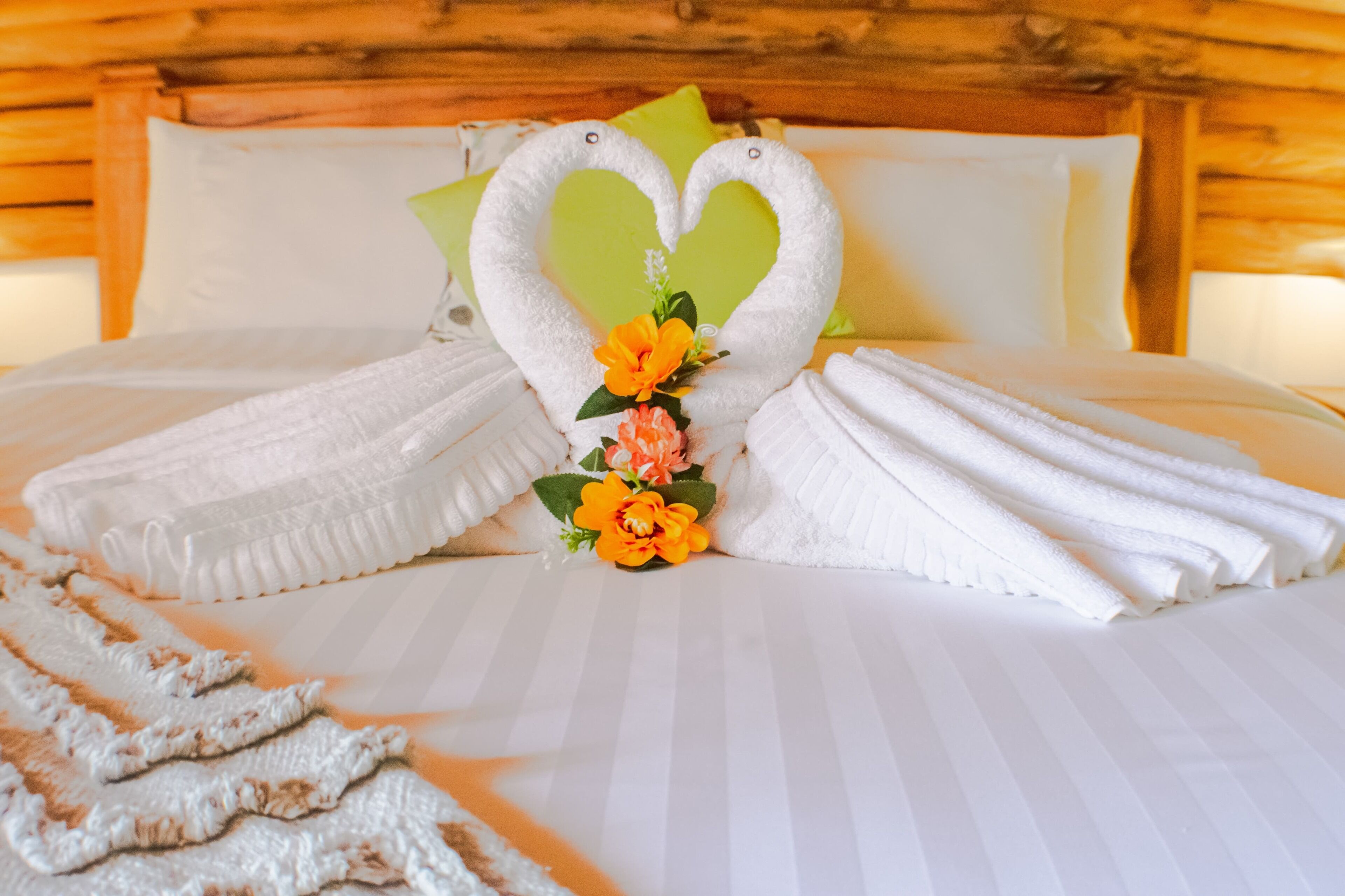 A neatly made bed with towels arranged in a heart shape, a small flower arrangement, and a wooden headboard.