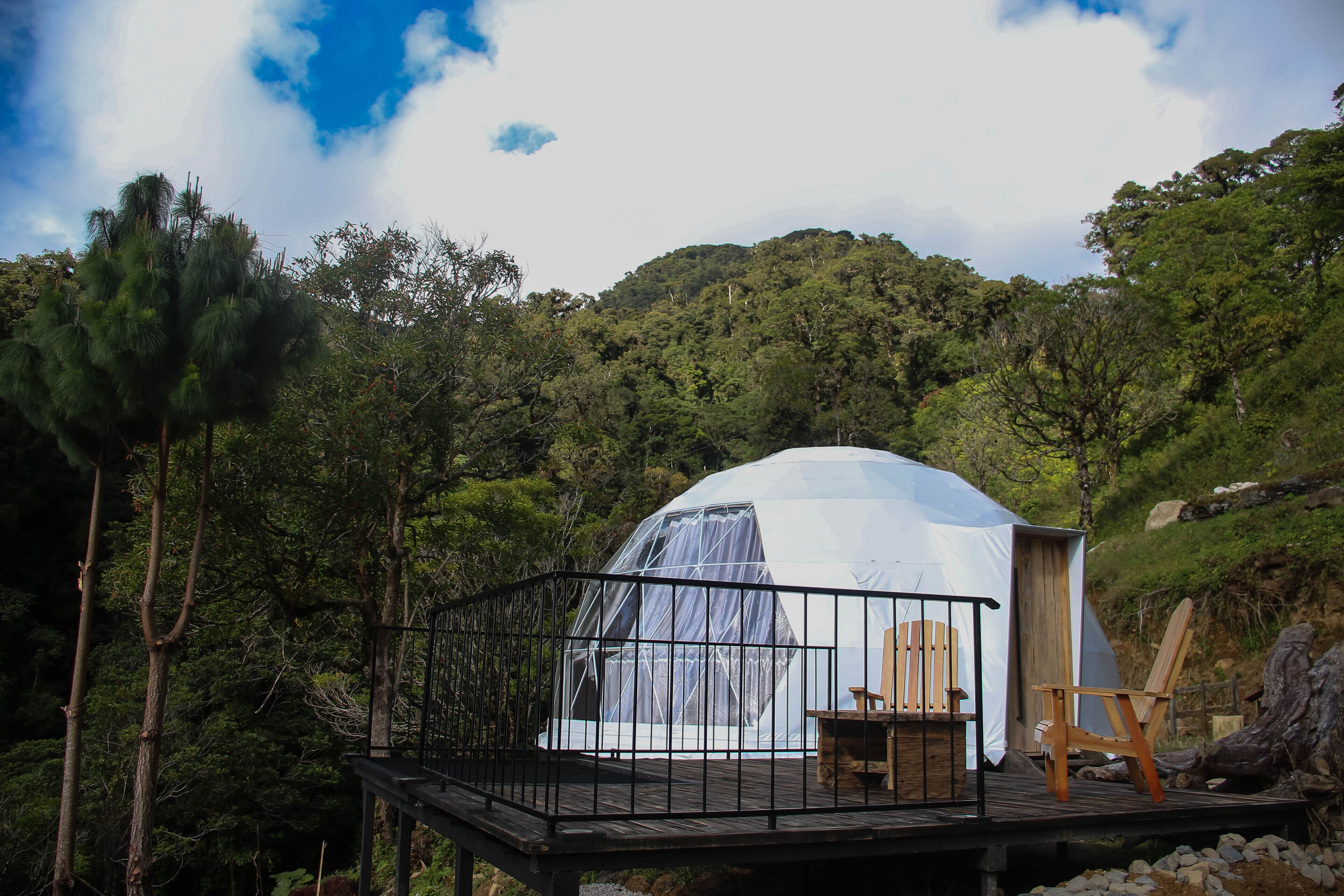 A dome-shaped tent with a wooden deck and two chairs, surrounded by dense forest and a clear sky.