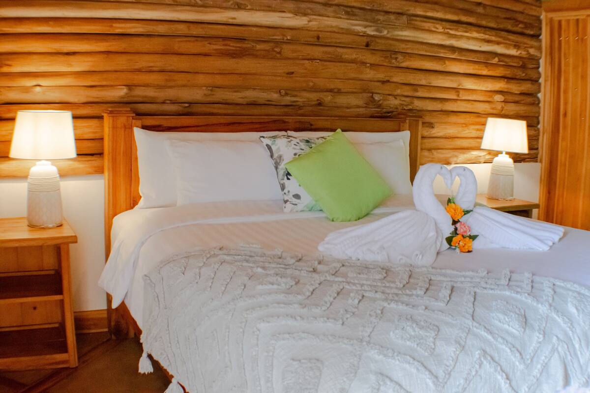 Una cama bien hecha con ropa blanca, una almohada verde y una decoración de toallas. Hay dos lámparas de noche sobre mesitas de madera.