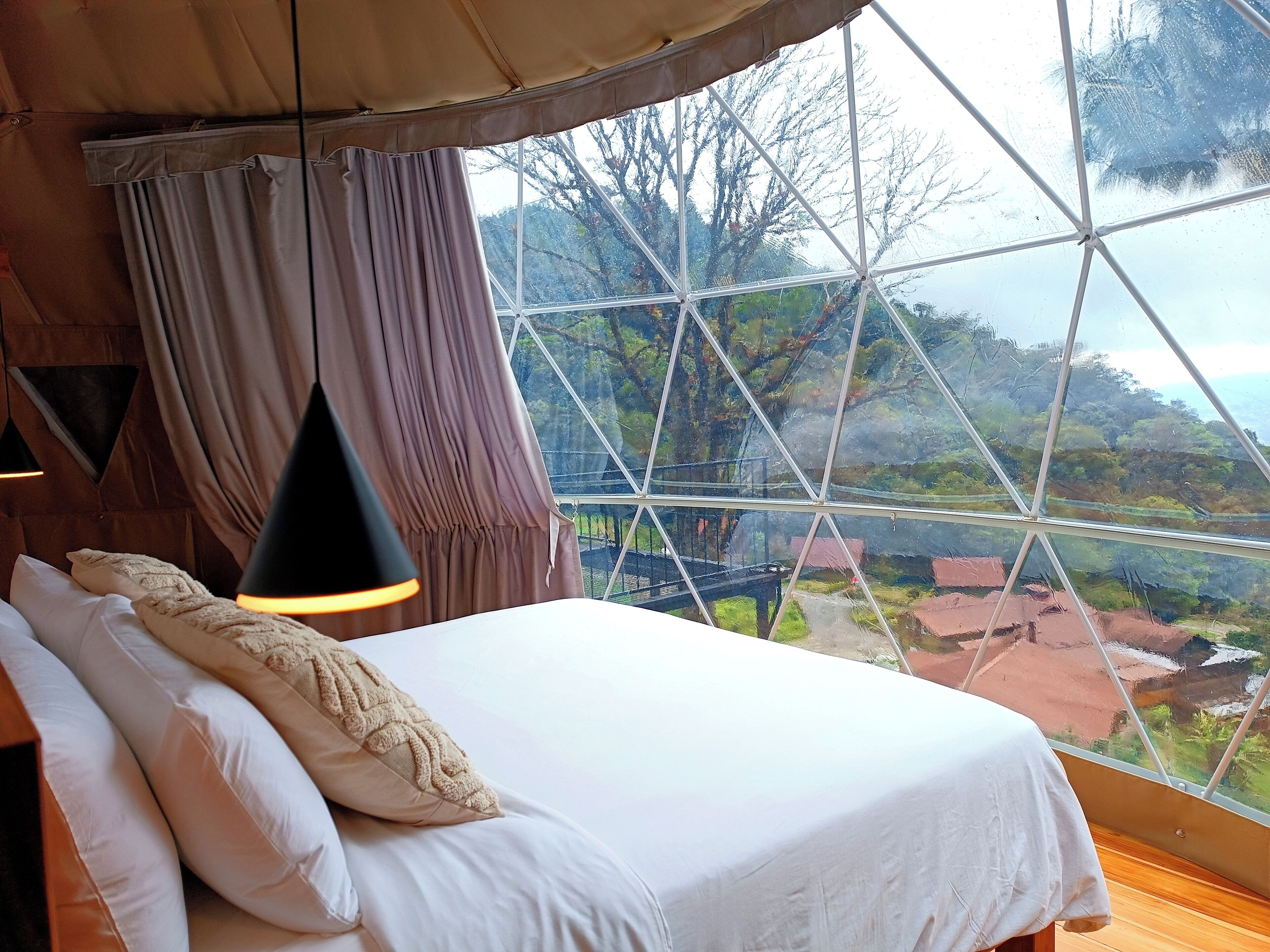 Ein Schlafzimmer mit großem Fenster, einem Bett mit weißer Bettwäsche, einer Hängelampe und Blick auf eine Landschaft mit Bäumen und Gebäuden.