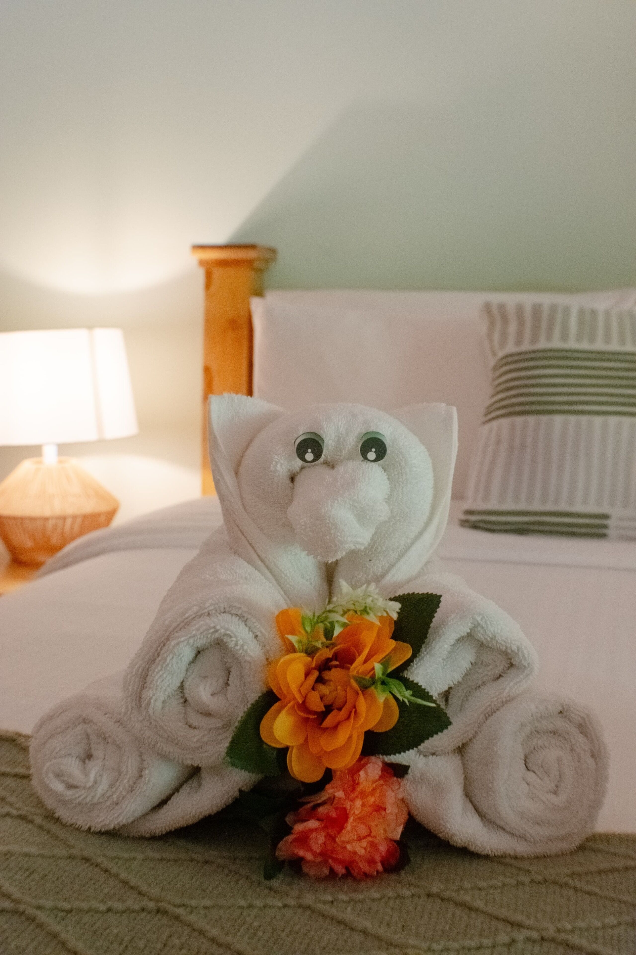 A neatly made towel sculpture of an elephant holding a flower, placed on a bed with a pillow and a lamp in the background.