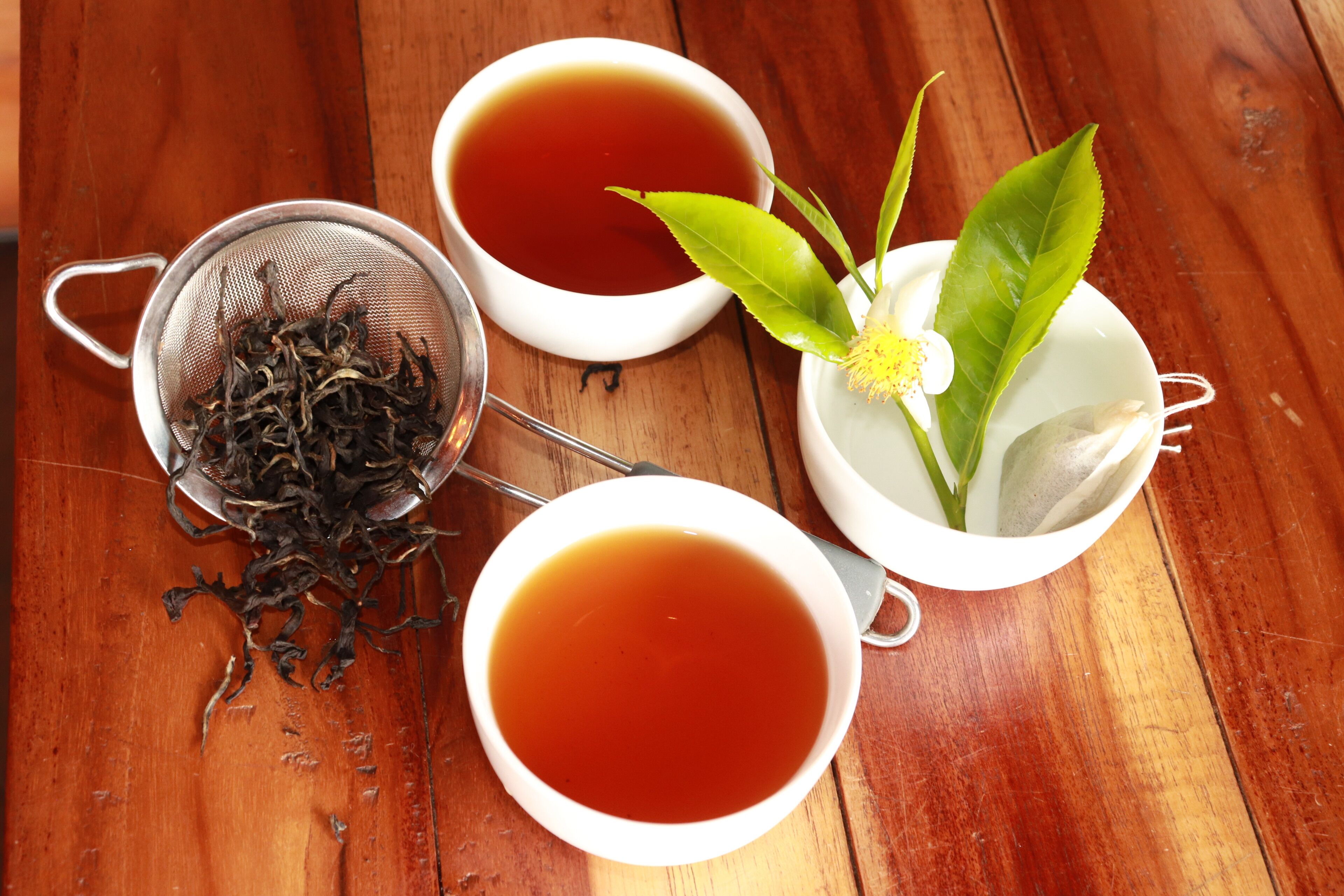 Three white cups of tea on a wooden table, one with a tea strainer and loose tea leaves, and another with a tea bag and a green leaf.