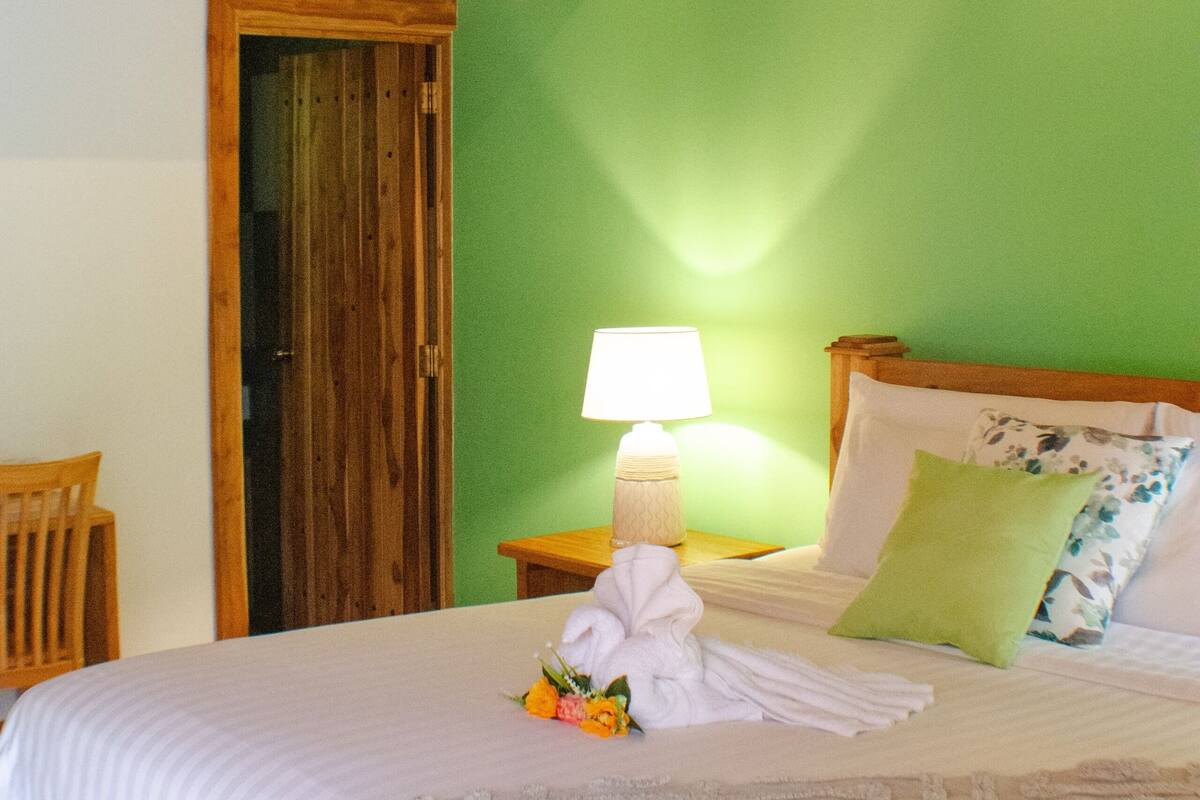 Un dormitorio con techo de madera, pared verde, cama con ropa blanca, mesita de noche con lámpara y puerta de madera.