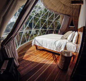 A bedroom with a bed, wooden flooring, a window with a view of trees, and a hanging light fixture. - Tree Trek Boquete Adventure Park (Los Naranjos)