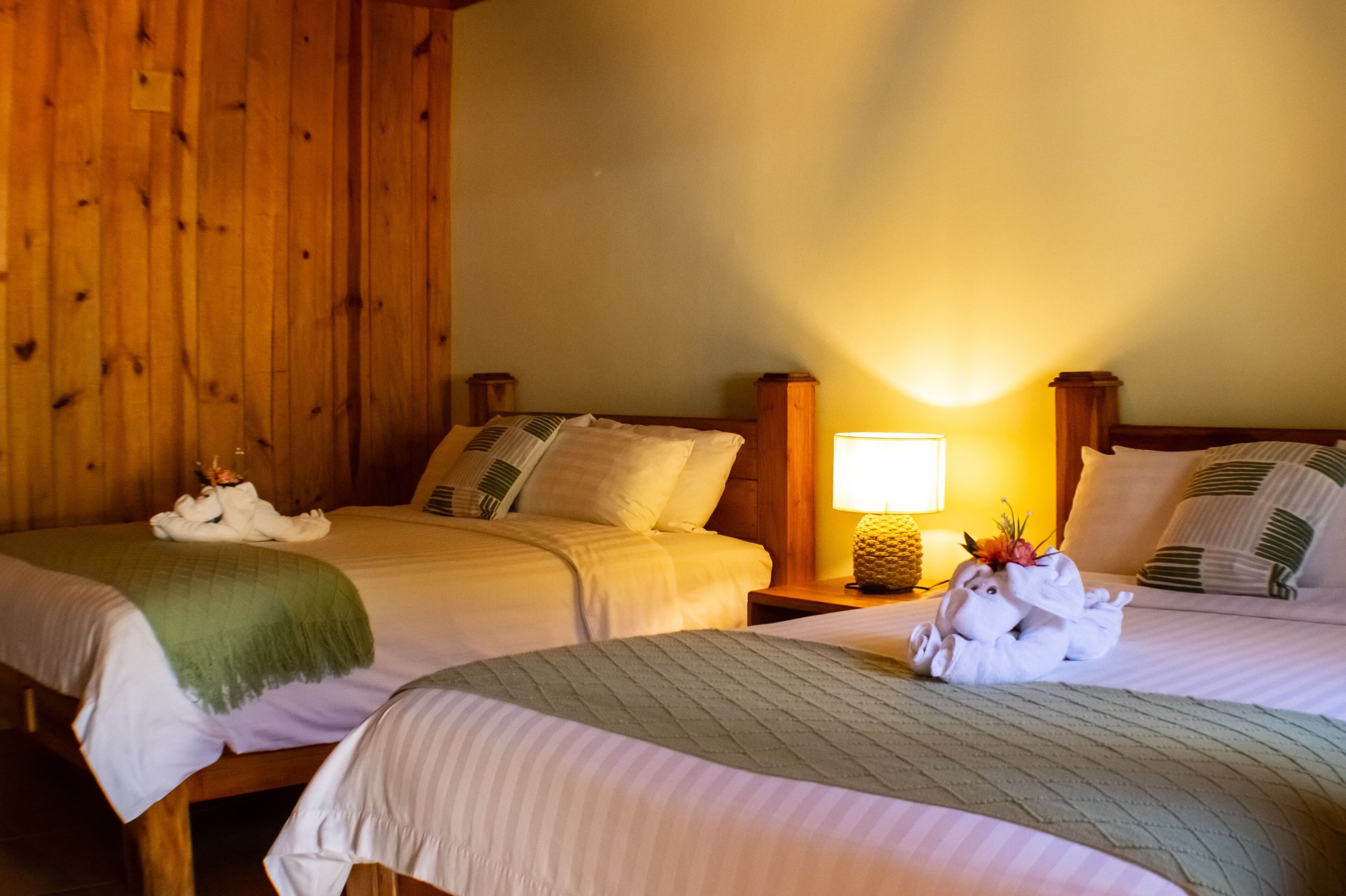 Two adjacent hotel beds with wooden headboards, a nightstand with a lamp, and a decorative pineapple.