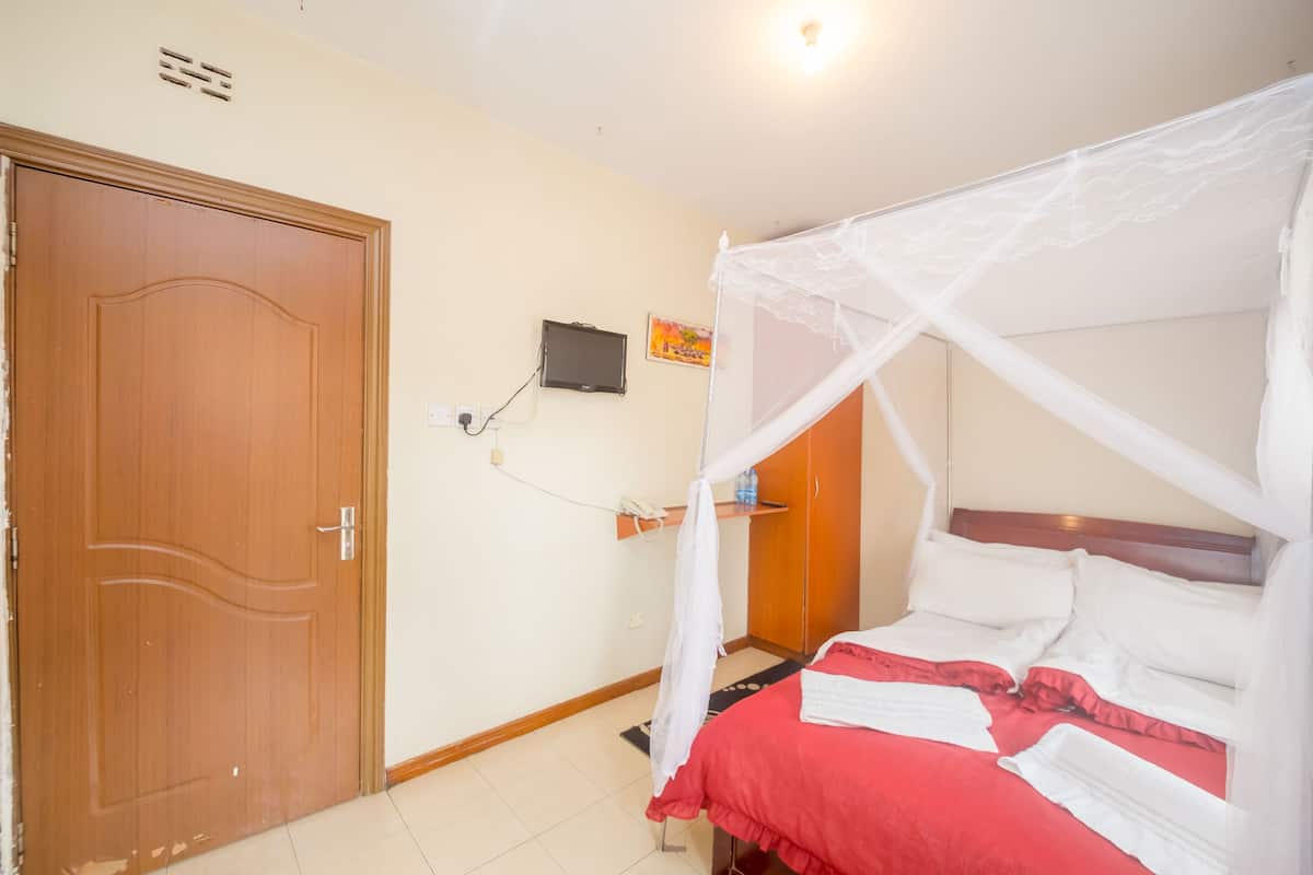 Een slaapkamer met een houten deur, een bed met een witte hemel, een rode spreideken en een klein plankje met een waterfles.