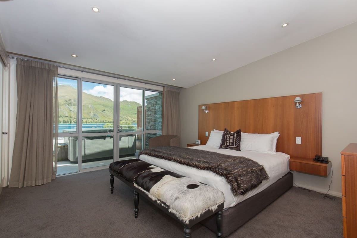 A spacious bedroom with a large bed, a bench, a wooden headboard, and a view of the outdoors through a sliding glass door.