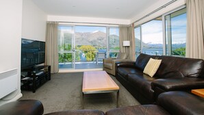 A modern living room with a leather sofa, a wooden coffee table, and a flat-screen TV. Large windows offer a view of mountains and a balcony. - Lakeside Apartments (Wanaka)