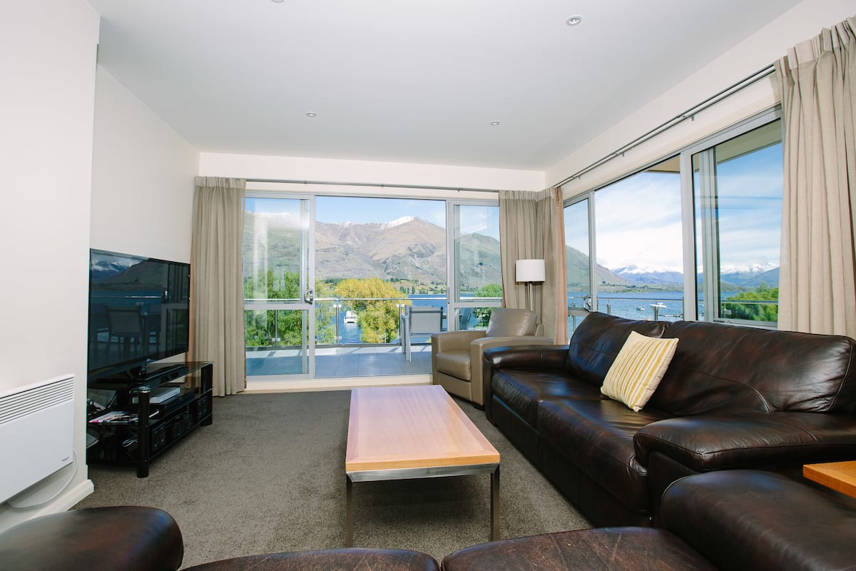 A modern living room with a leather sofa, a wooden coffee table, and a flat-screen TV. Large windows offer a view of mountains and a balcony.