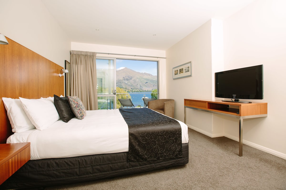 A hotel room with a large bed, a TV, a desk, and a view of a mountain and lake.