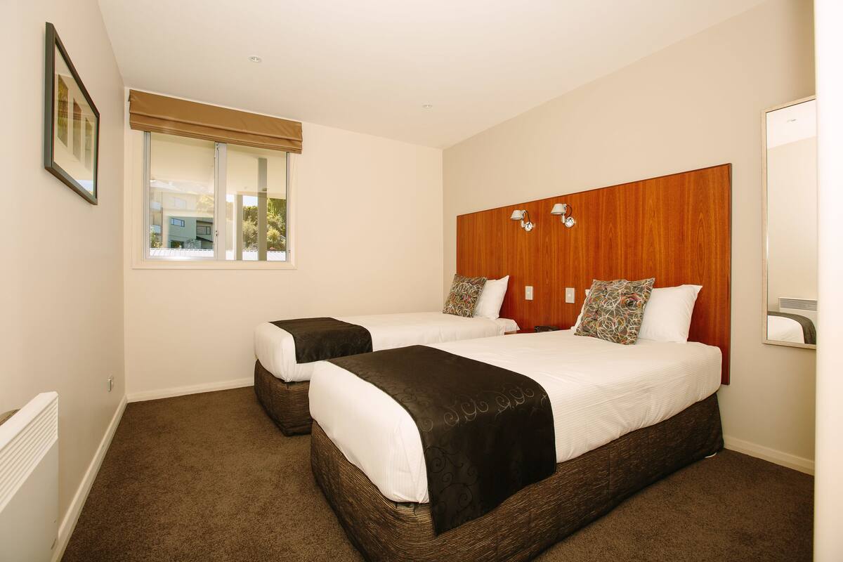 A hotel room with two single beds, a large window with blinds, a wooden headboard, and a mirror on the wall.