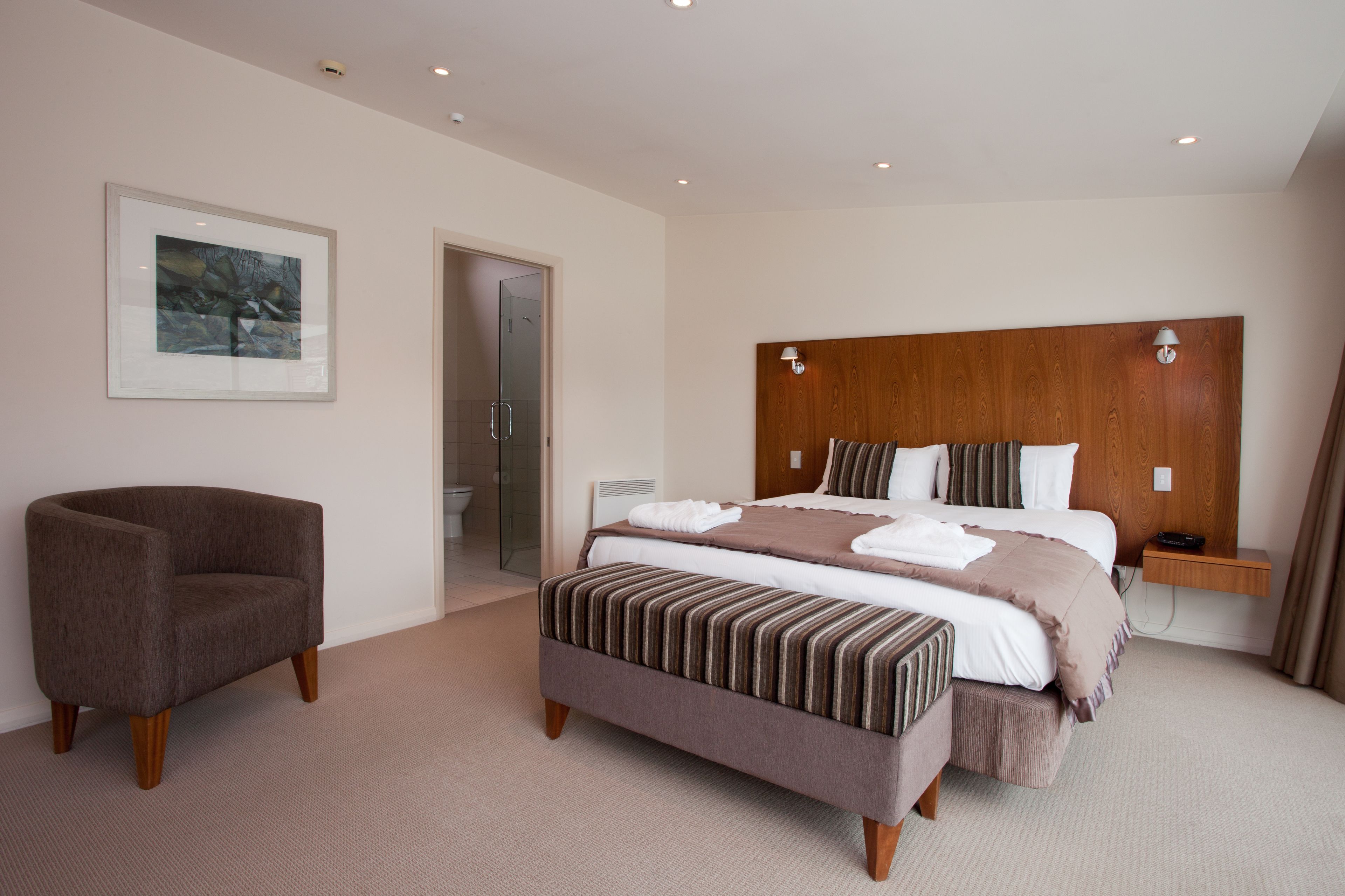 Une chambre d’hôtel moderne avec un grand lit, une tête de lit en bois, une table de chevet, une chaise et un tableau encadré au mur.