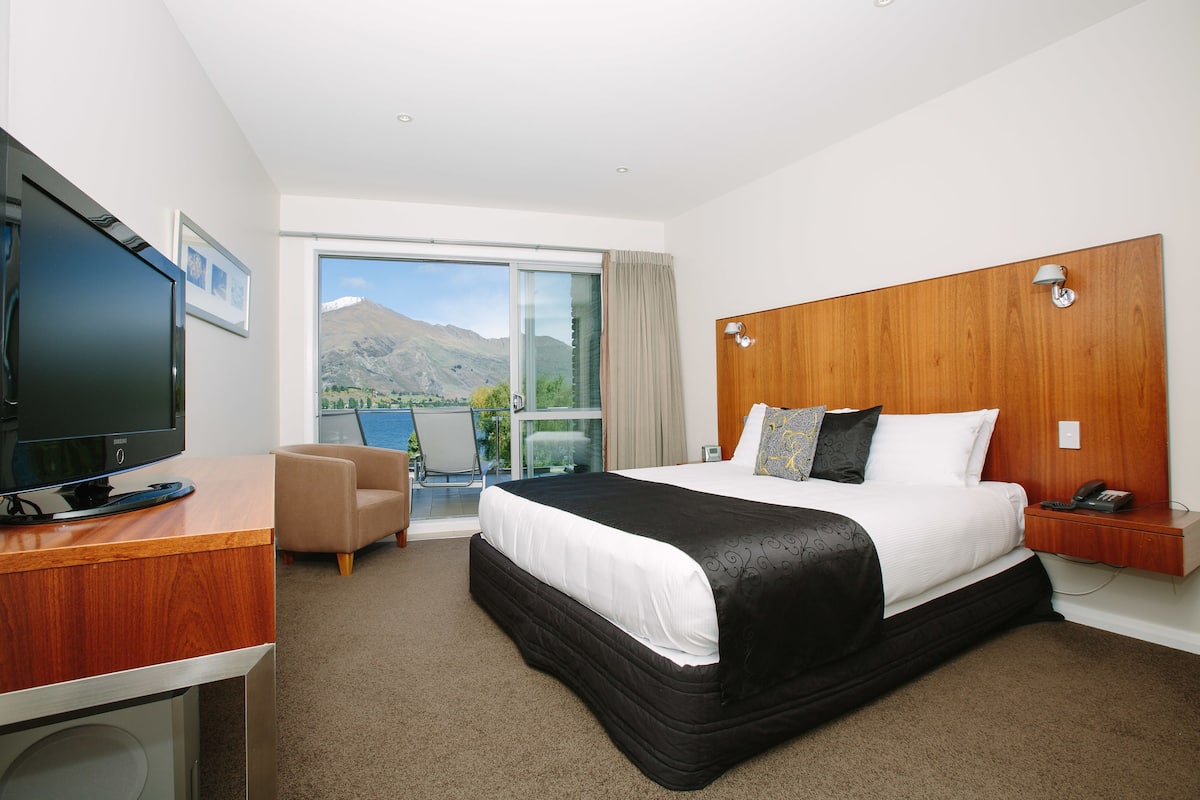 A hotel room with a bed, a desk with a TV, a chair, a small table, and a view of mountains through a large window.
