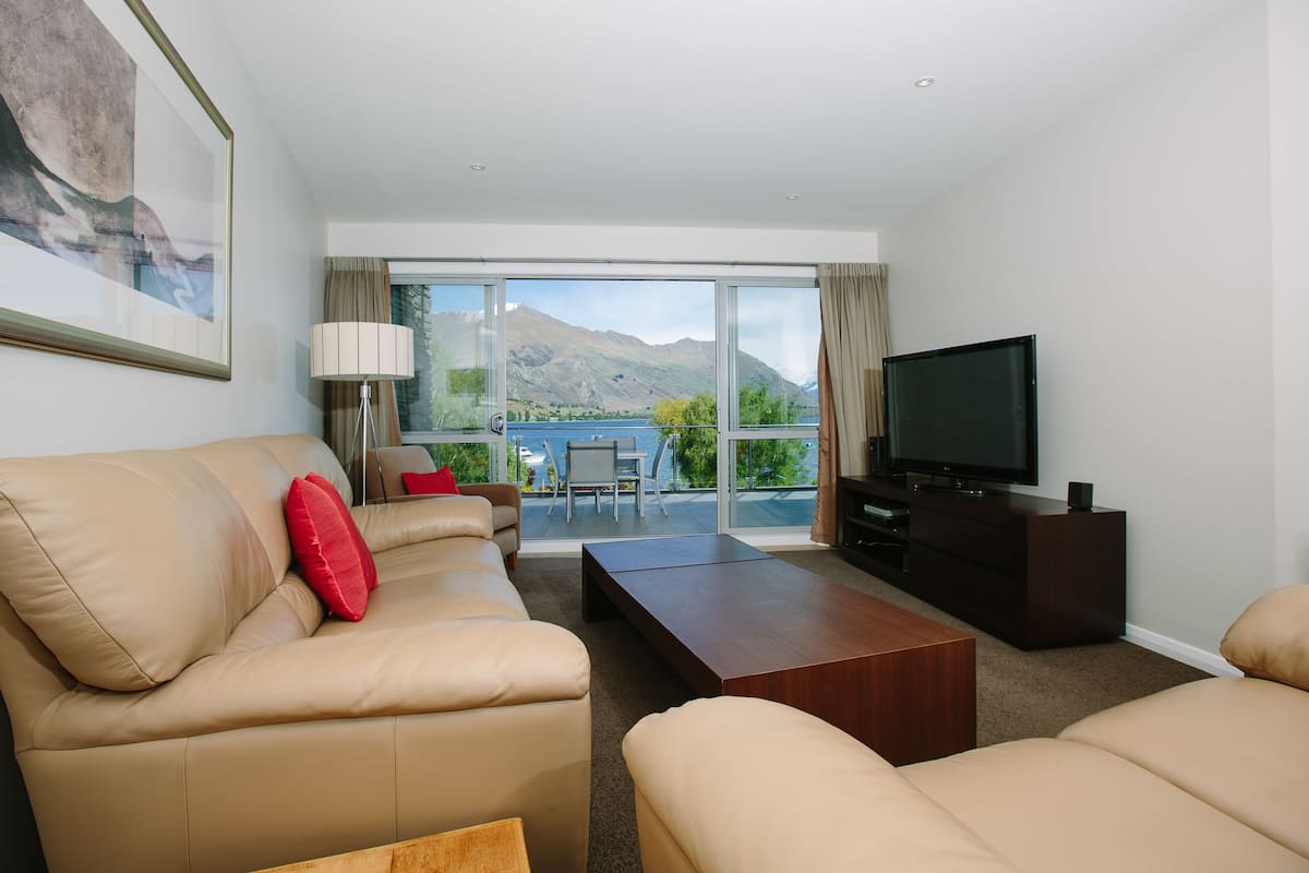 A modern living room with a sofa, a coffee table, and a TV. There is a large window with a mountain view.