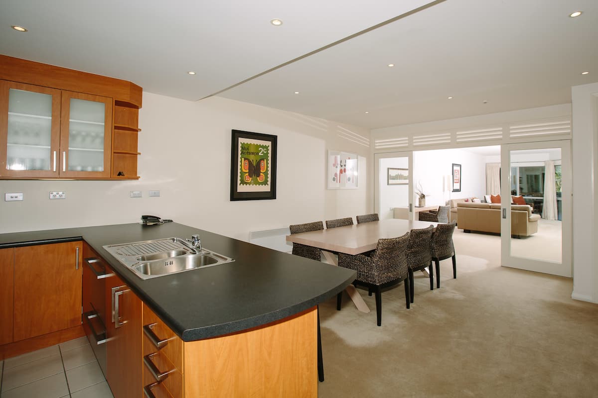 A modern kitchen with a central island, dining area with wicker chairs, and a living room with a sofa and a large window.