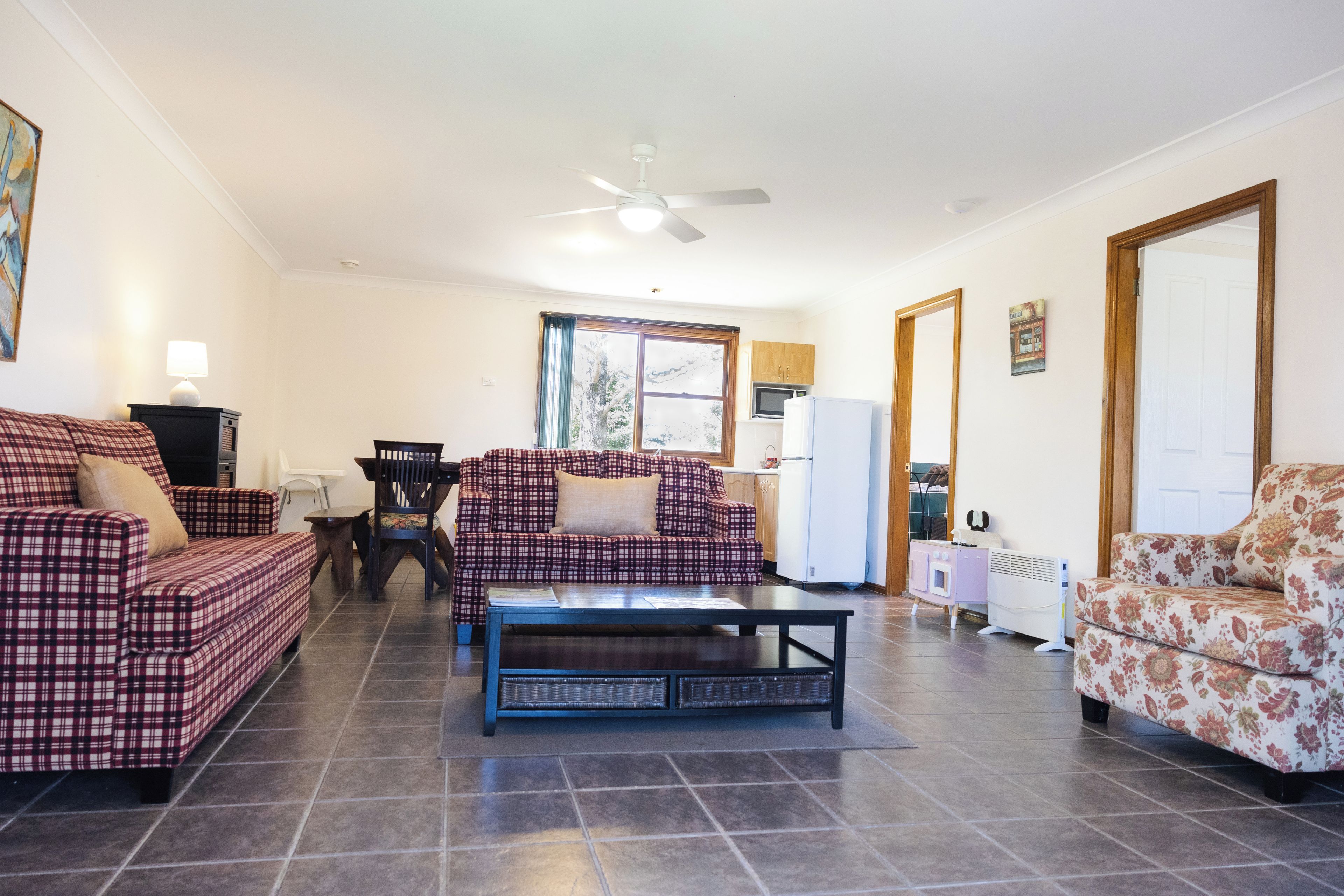 Un salon comprenant un canapé, un fauteuil, une table basse, un ventilateur de plafond et un coin cuisine avec un réfrigérateur.