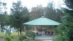 Un quiosco con techo verde y una mesa de picnic en el centro, rodeado de árboles y un césped.