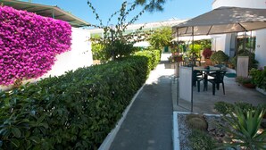 Un sendero bordeado por flores moradas y setos verdes, que conduce a un área de estar al aire libre con una mesa y sillas bajo una pérgola.