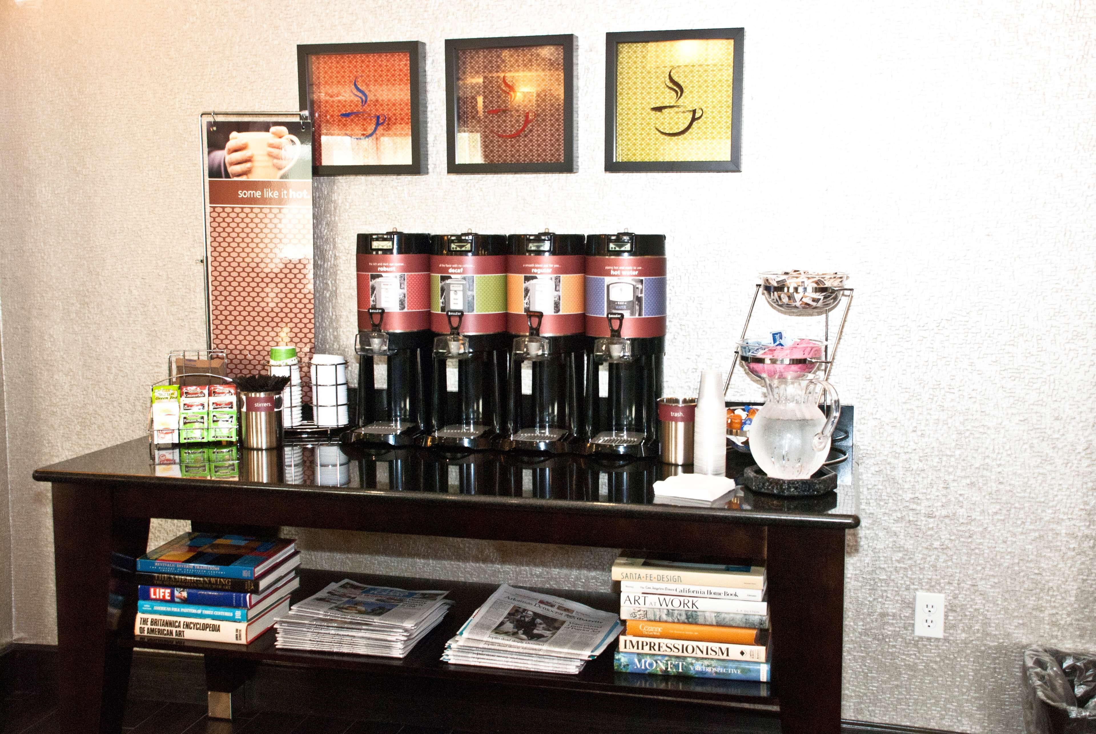 A coffee station with multiple coffee dispensers, a water pitcher, and a basket. There are books and magazines on a table below.