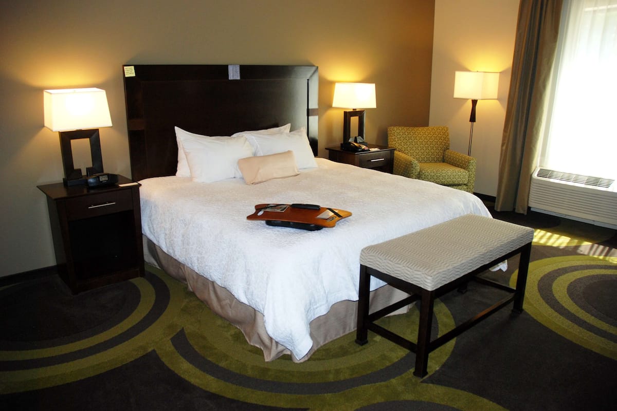 A hotel room with a large bed, two bedside tables with lamps, a chair, a bench, and a carpeted floor.