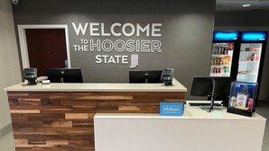 Un comptoir d’accueil d’hôtel avec une pancarte indiquant « Bienvenue dans l’État de Hoosier », une autre pancarte « Hilton Honors » et un distributeur de boissons.