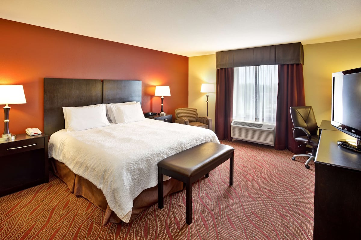 Une chambre d’hôtel avec un grand lit, deux tables de chevet avec des lampes, un banc, un bureau avec une télévision et une fenêtre avec des rideaux.