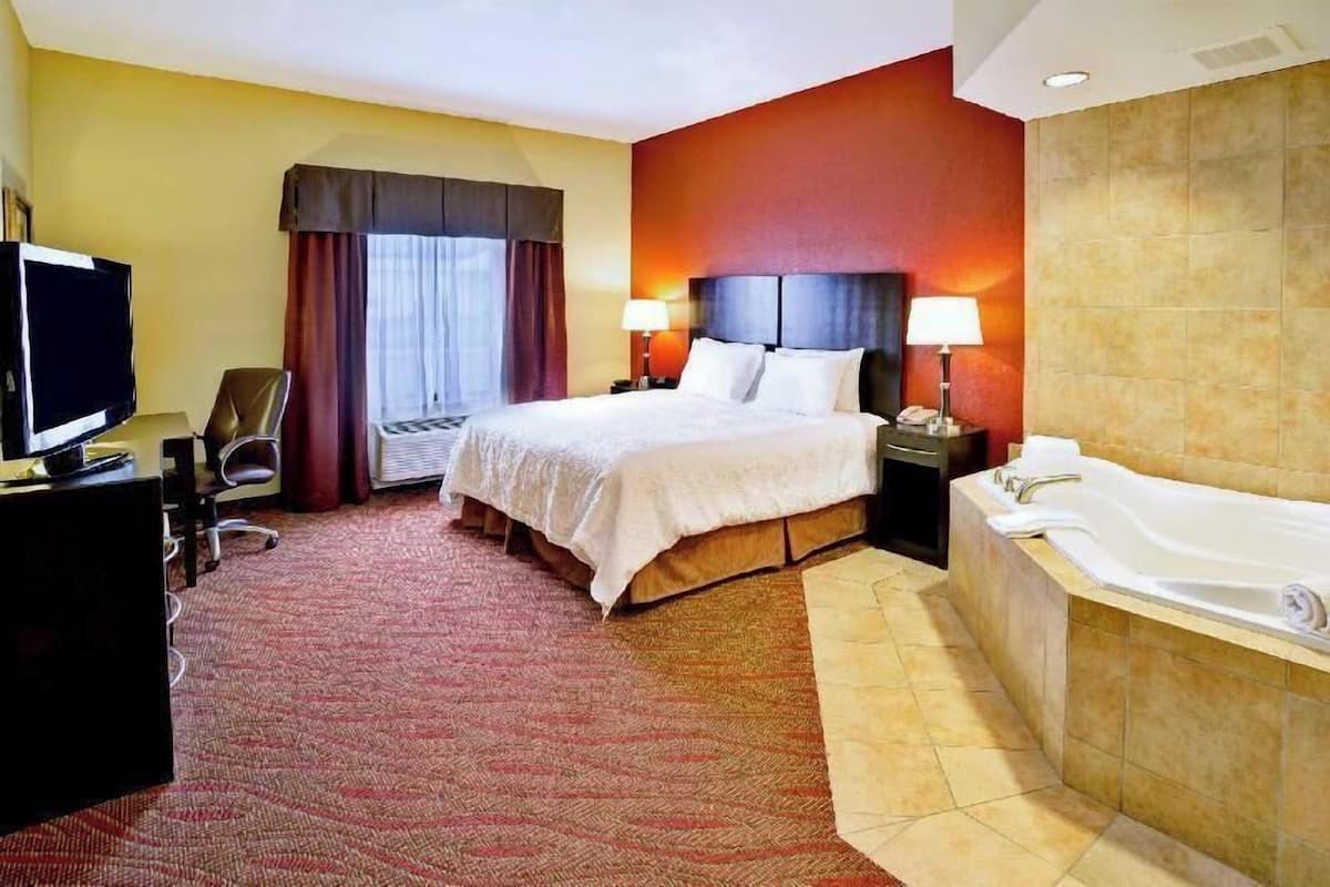 Une chambre d’hôtel comprenant un lit, un bureau avec une télévision, une chaise, une salle de bain avec une baignoire et un mur d’accent de couleur rouge.