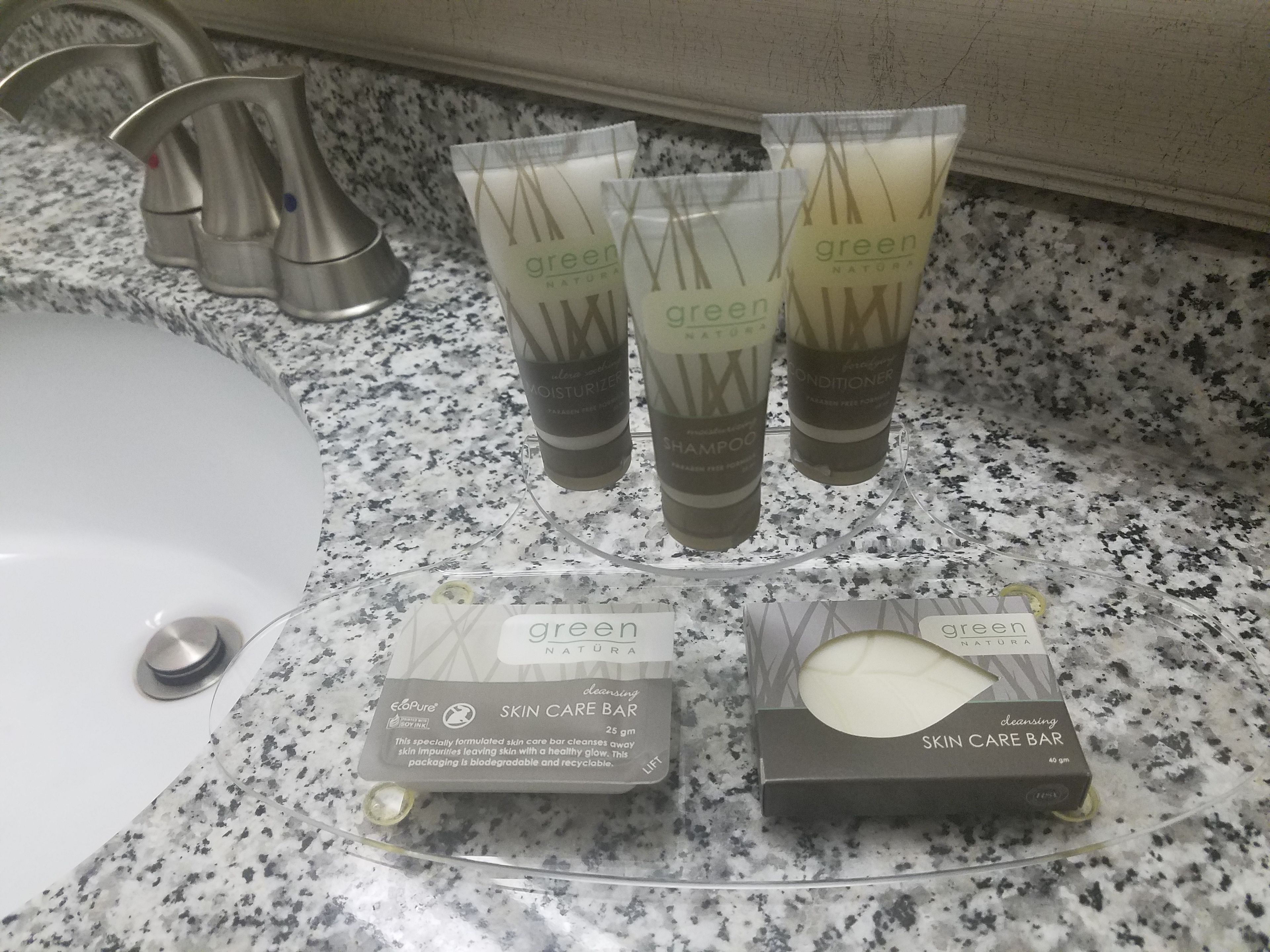 A bathroom counter with a granite sink, a silver faucet, and three tubes of green Naturia hair care products. Below the tubes, there are two boxes of green Naturia skin care bar products.