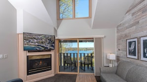 A living room with a fireplace, a television, a sofa, and a balcony with a view of a lake. - Country House Resort (Sister Bay)