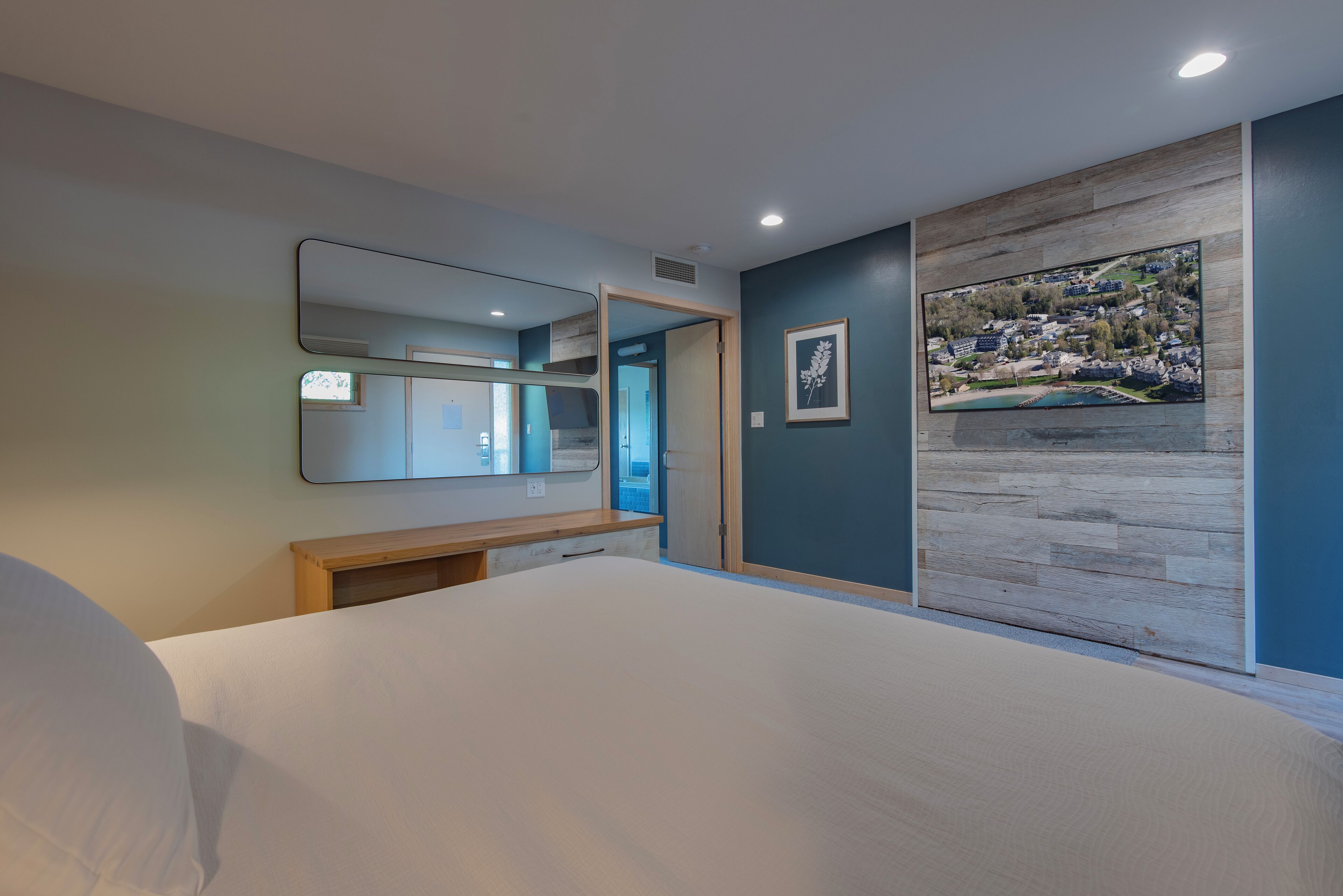 A bedroom with a large bed, a wooden bedside table, a mirror, and a view of a town through a large window.