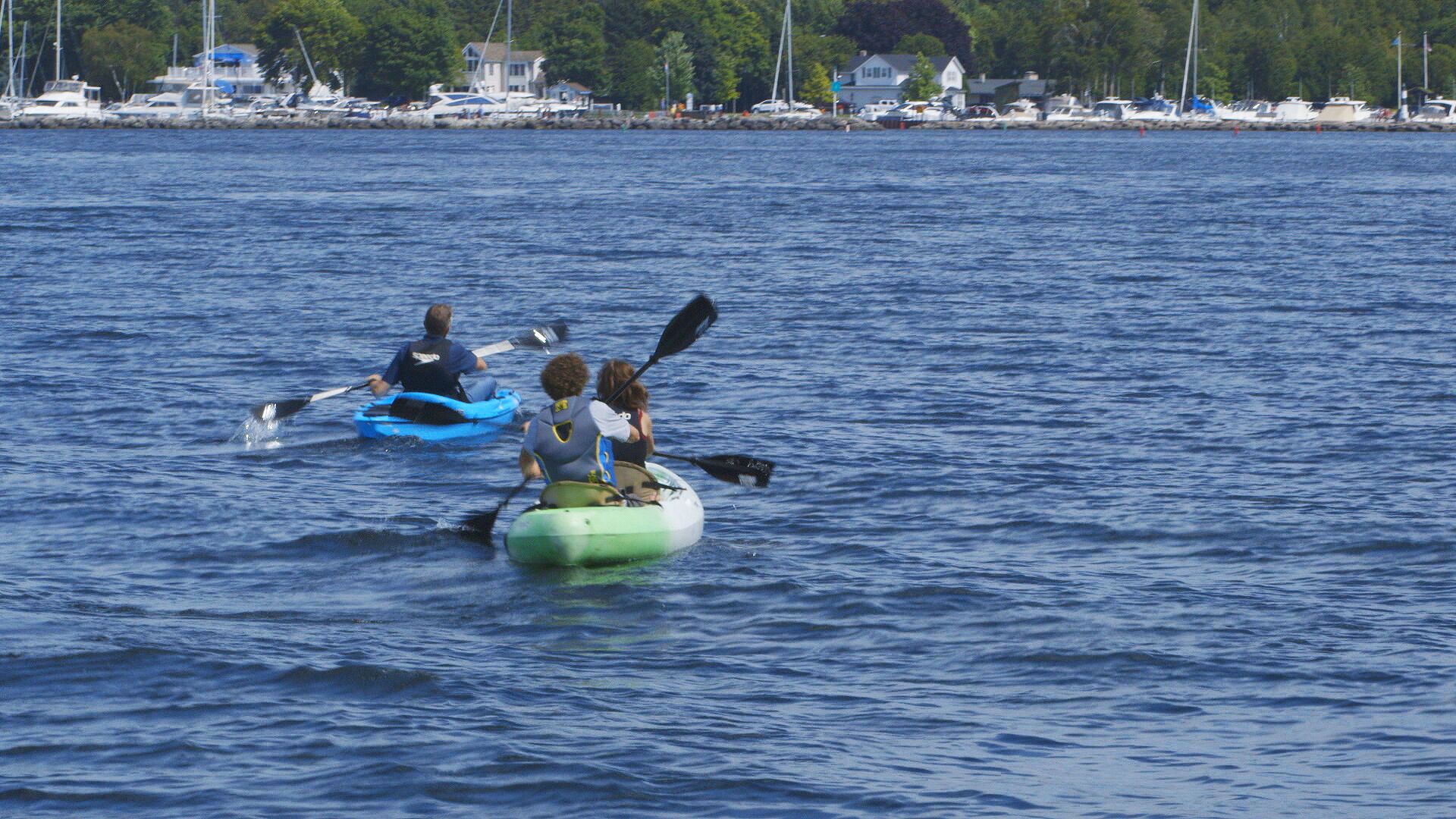 Three kayaks on a calm body of water with a marina and houses in the background.
