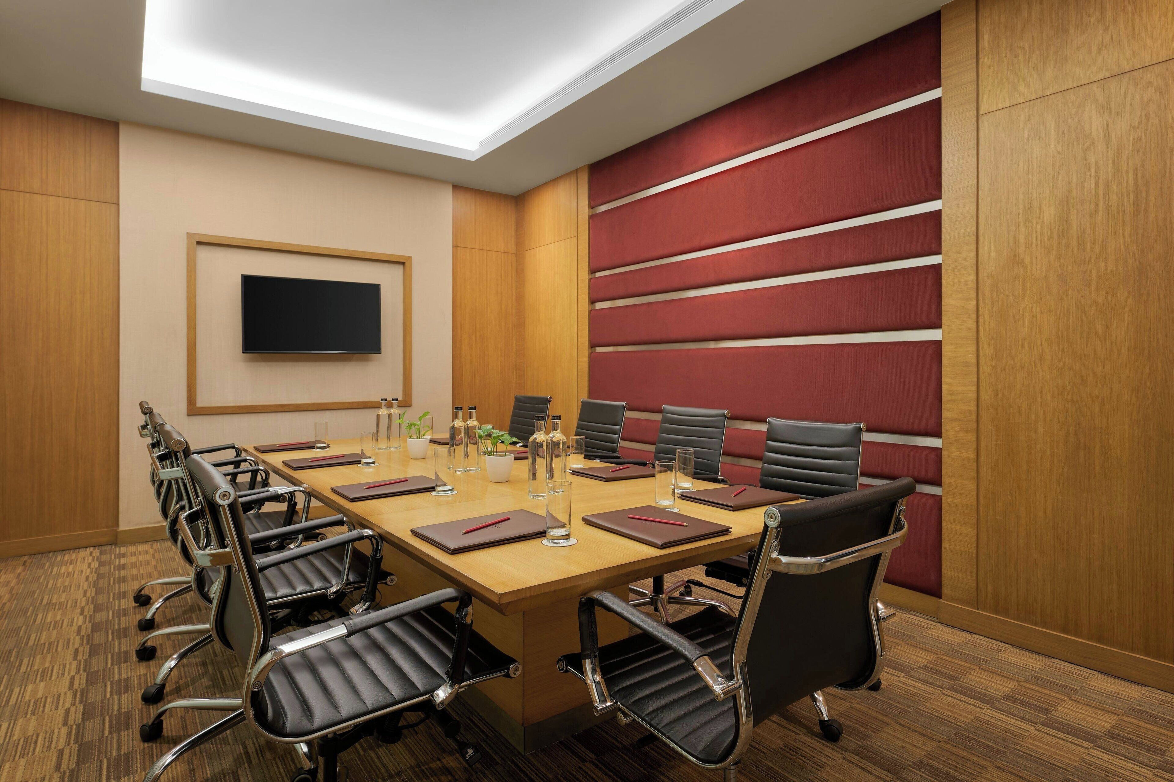 A conference room with a long table, multiple chairs, a flat-screen TV, and a red accent wall.