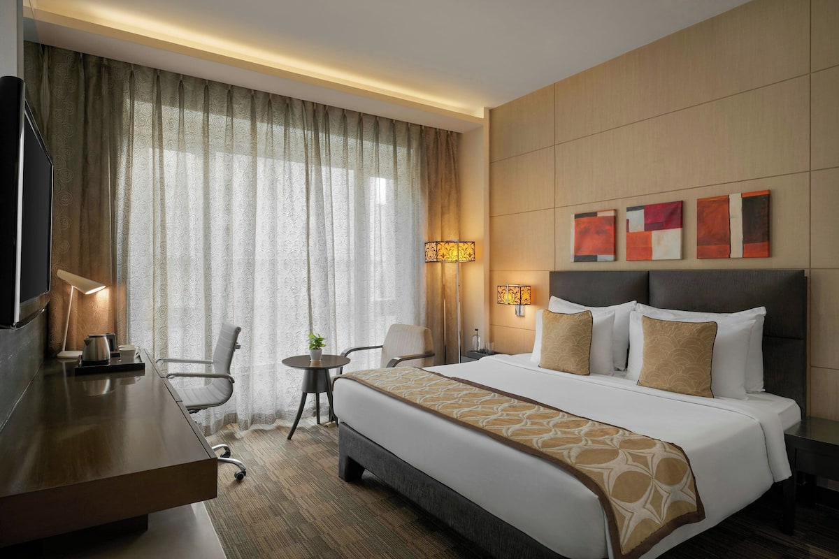 A hotel room with a bed, a desk, a chair, a small table with a plant, and a TV.