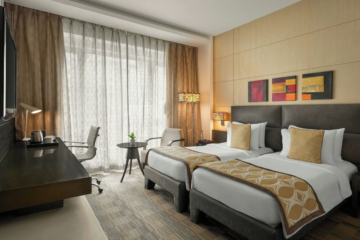 A hotel room with two beds, a desk, a chair, a small table, and a lamp.