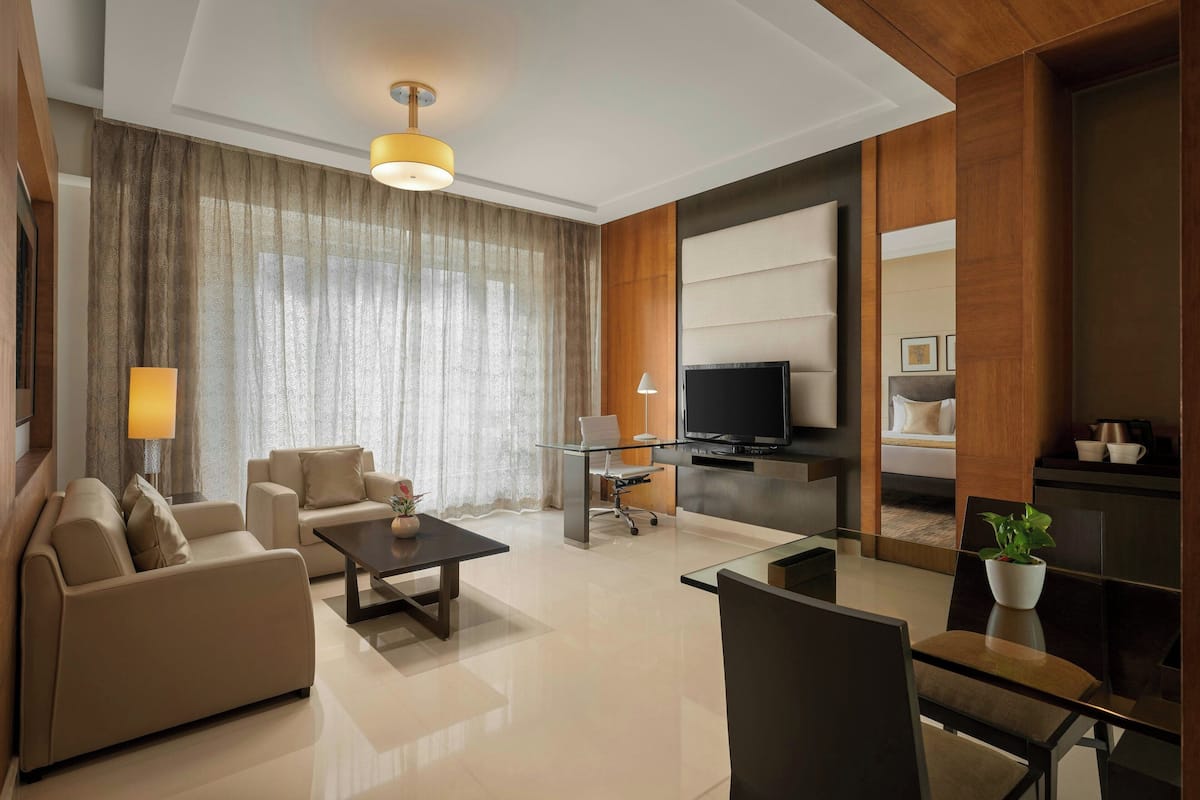 A modern hotel room with a sofa, a glass coffee table, a TV, and a dining area.