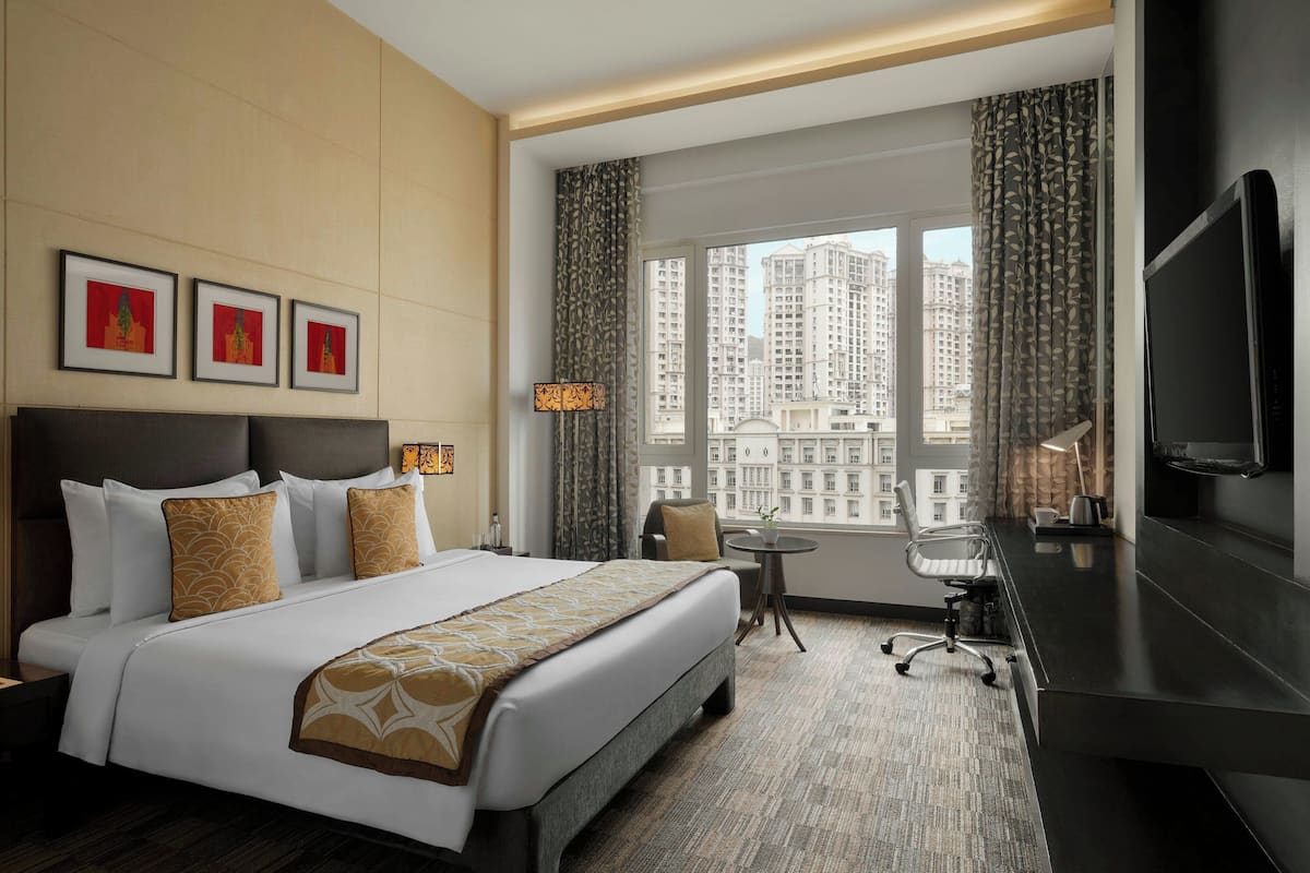 A hotel room with a large bed, a desk, a chair, a TV, and a view of tall buildings.