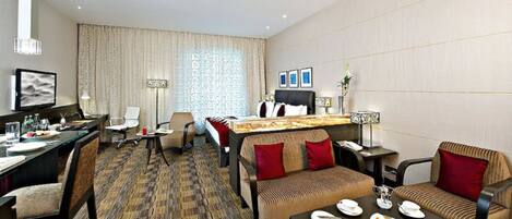 A hotel room with a bed, a desk with a computer, a sofa, a dining table with plates and glasses, and a large window with curtains.