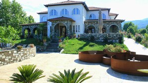 A large, multi-story house with a stone facade, arched windows, and a well-maintained garden with planters and palm trees. - Zeus Hotel (Dio-Olympos)