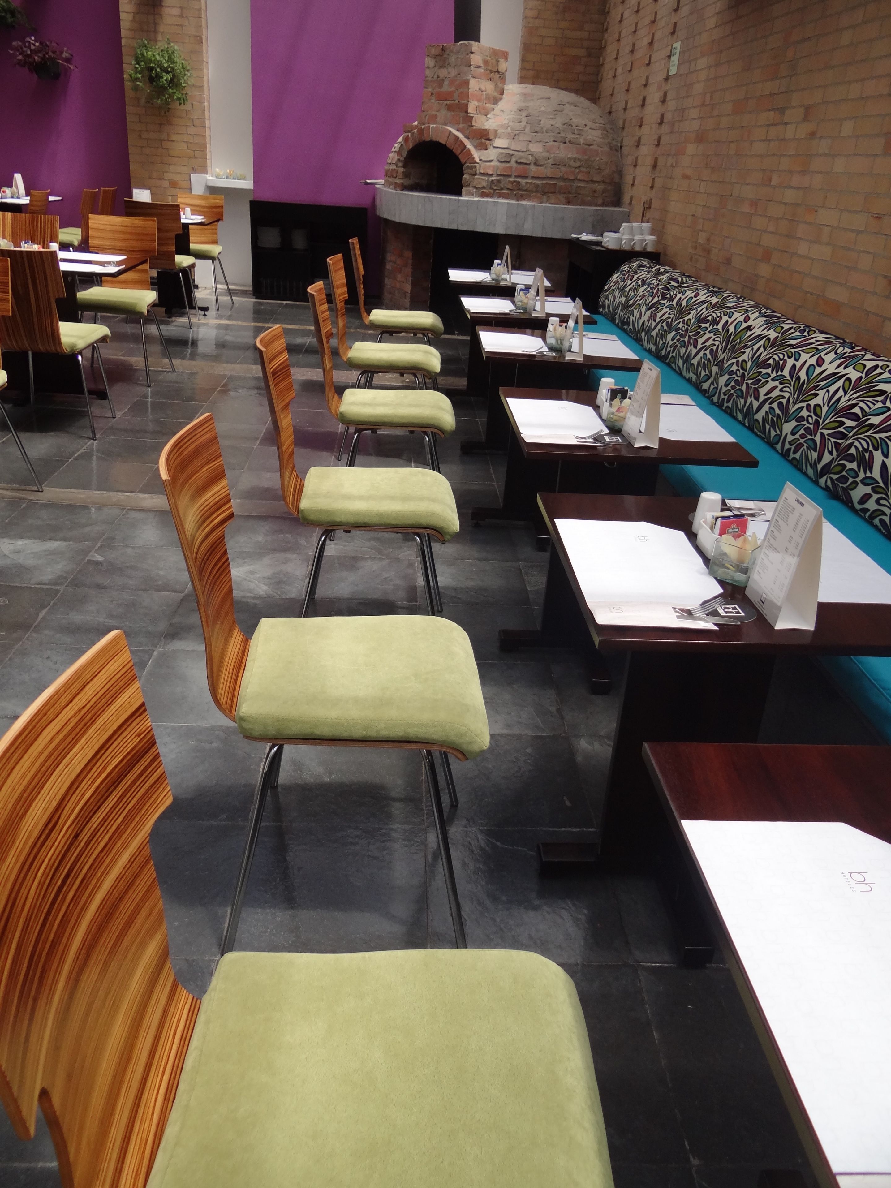 A restaurant with a brick oven, green cushioned chairs, and a long communal table.