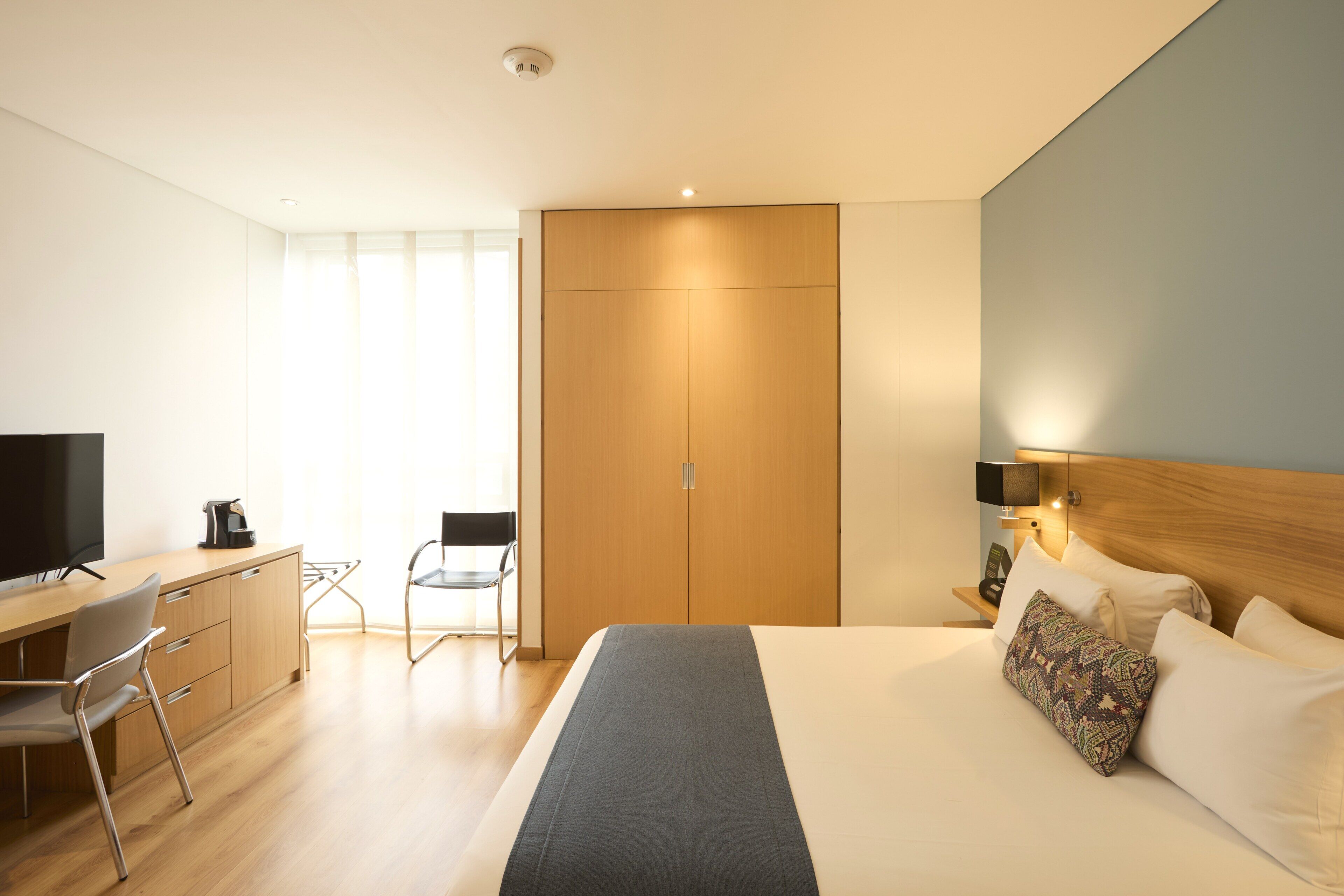A hotel room with a bed, a desk with a chair, a television, and a wardrobe.