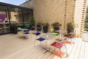 Un patio exterior con mesas y sillas de metal, rodeado de macetas con plantas y un muro de ladrillo.