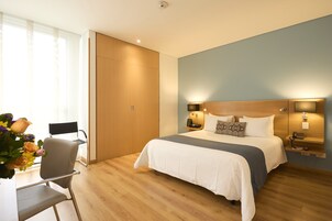 Habitación de hotel moderna con piso de madera, cama con ropa blanca, escritorio con silla y un gran ventanal con cortinas.