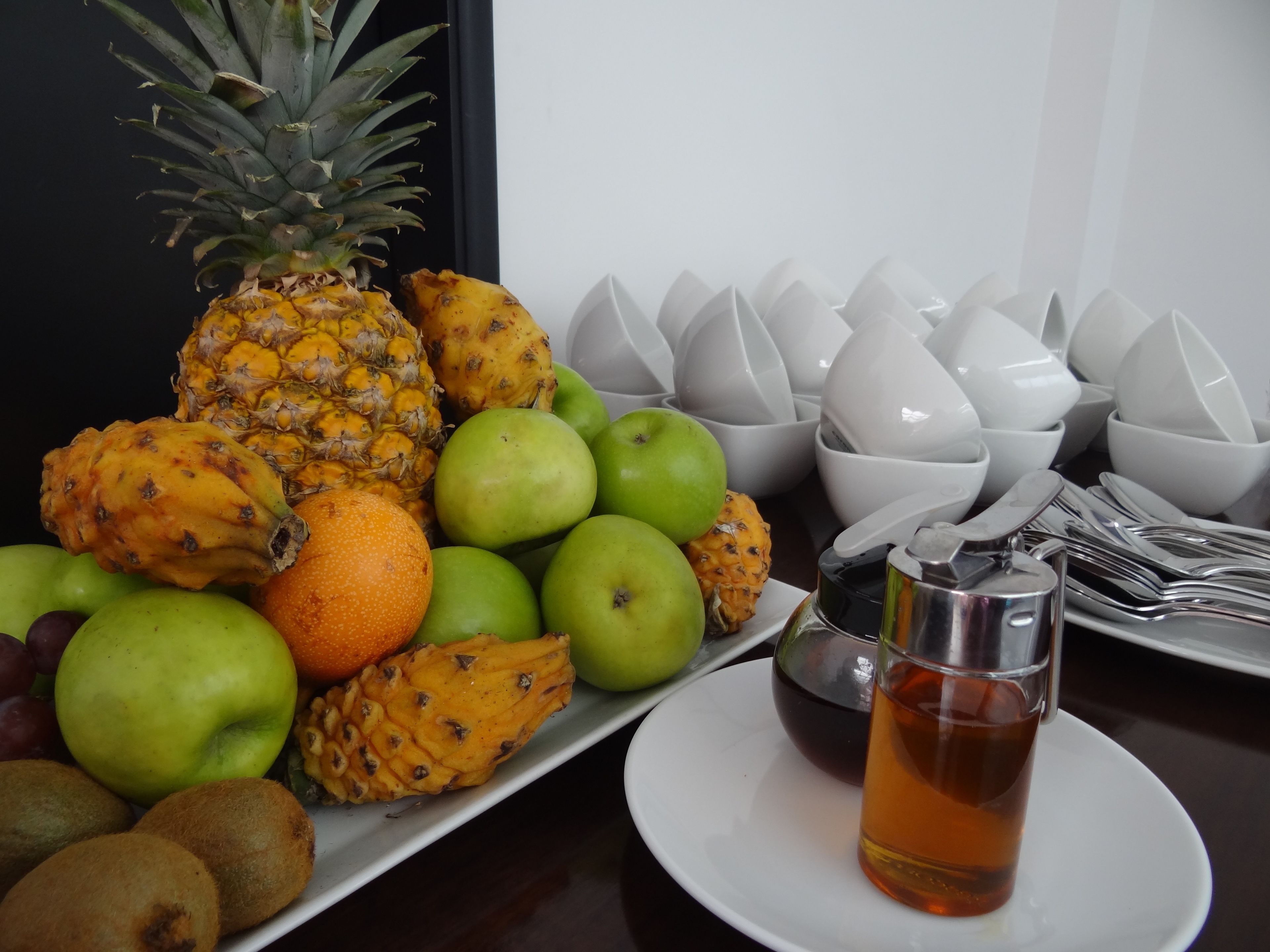 Un vassoio di frutta con ananas, mele e arance, un bicchiere di tè e ciotole bianche su un tavolo.