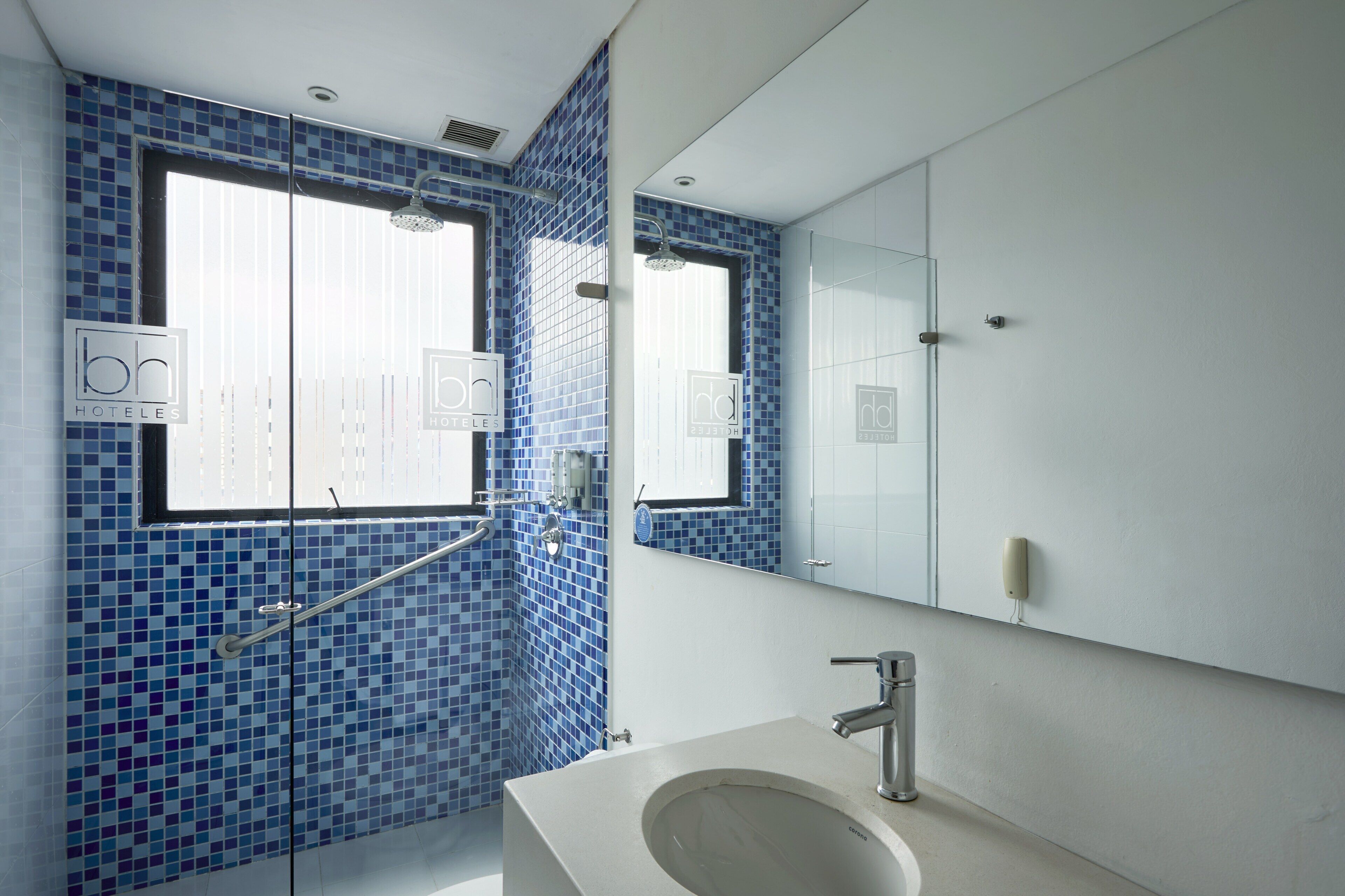 A modern bathroom with blue mosaic tiles, a large mirror, a sink with a faucet, and a window with a sign.