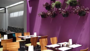 Un espace repas aux murs violets, avec plusieurs tables et chaises. Des plantes en pot sont disposées le long du mur, et un coin salon est aménagé avec un canapé à motifs.