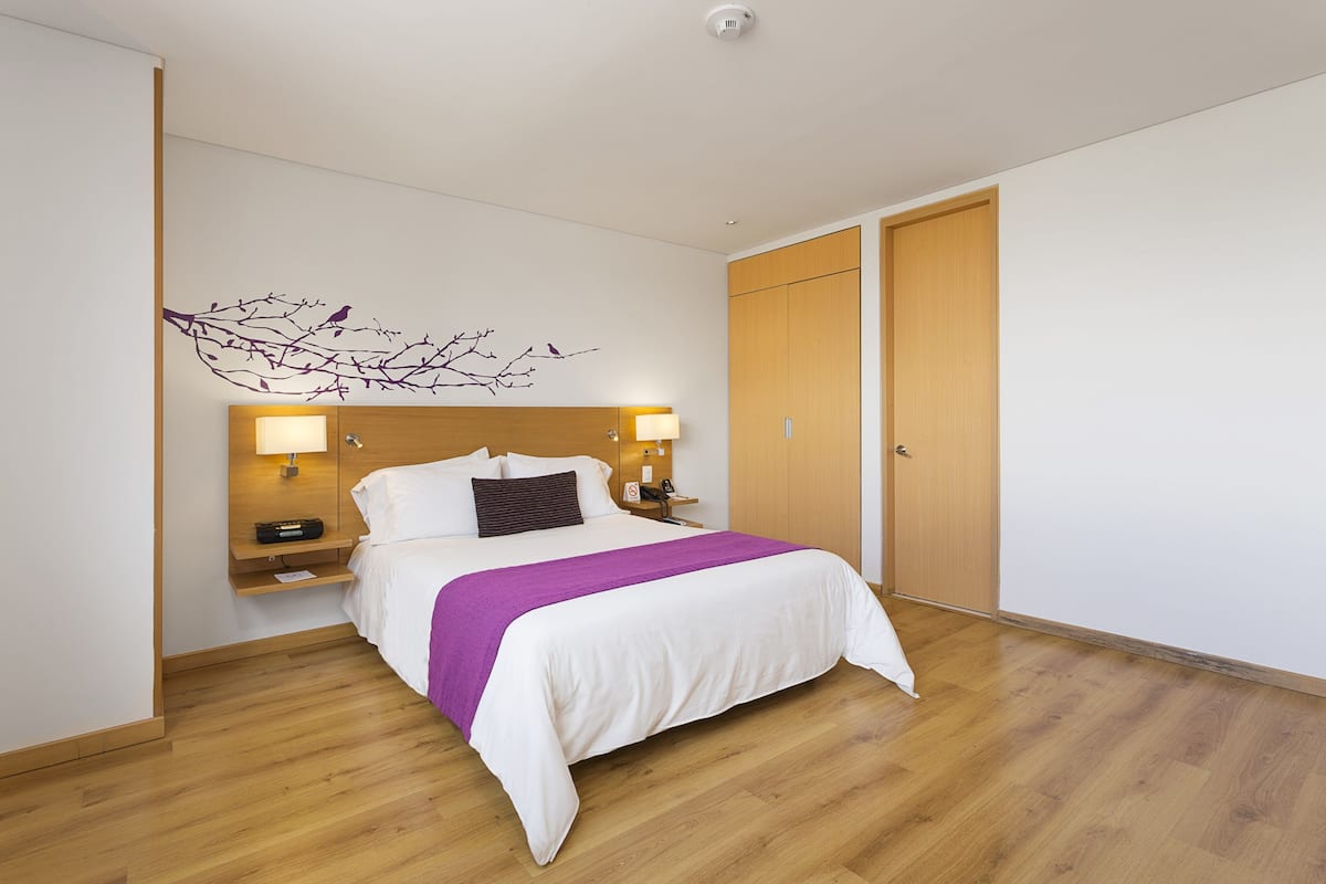 A hotel room with a bed, wooden floor, a wall-mounted branch decoration, a nightstand with a lamp, and a door.