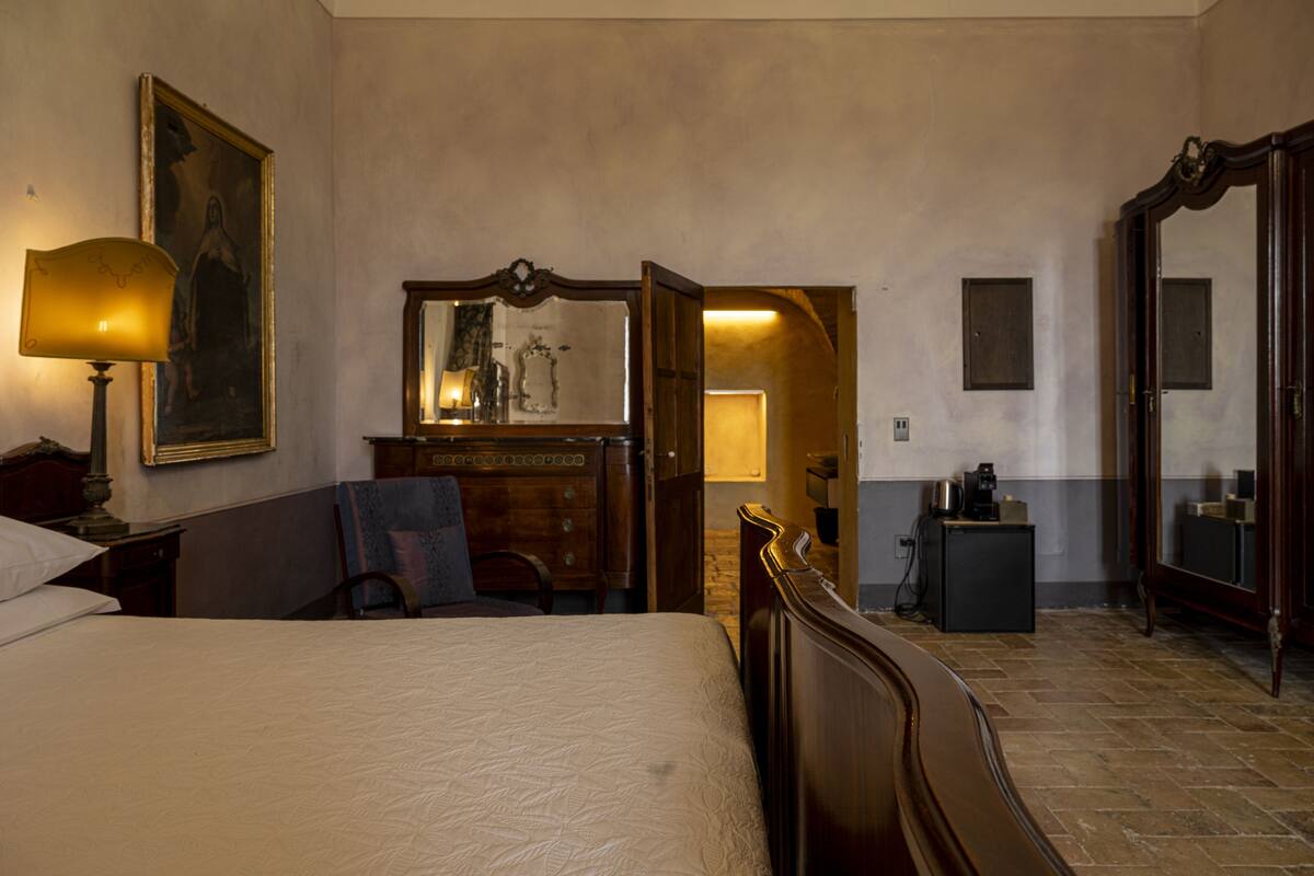 A hotel room with a large bed, a wooden dresser, a desk with a lamp, a mirror, and a door leading to another room.