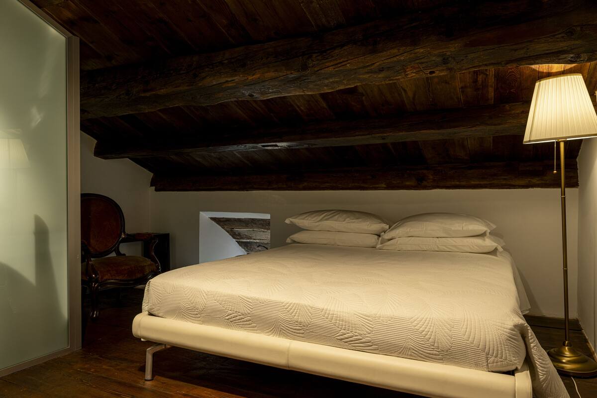 A bedroom with a wooden ceiling, a bed with a white bedspread, a bedside lamp, and a chair.