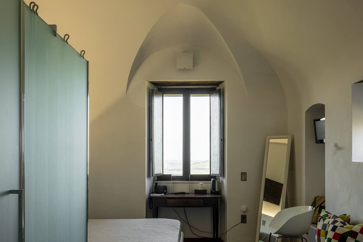 A bedroom with a bed, a desk, a chair, a mirror, and a window.