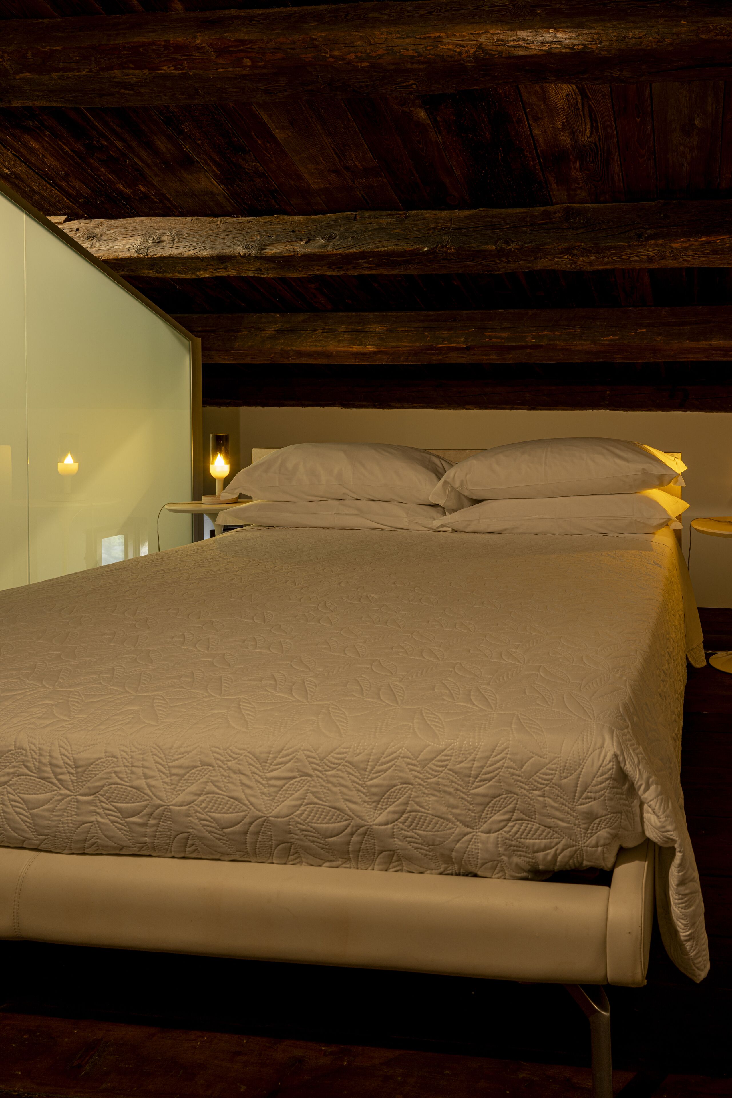 Uma cama com cobertura acolchoada, vigas de madeira no teto e abajures laterais.